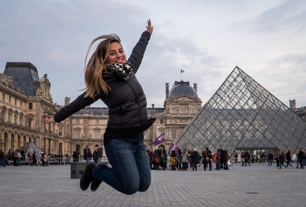 Kate jumping in the air, her knees tucked beneath her, in front of the Louvre Museum in Paris and it's glass pyramid.