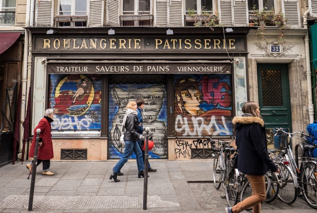Two women walking past a Boulangerie Patisserie covered in graffiti.