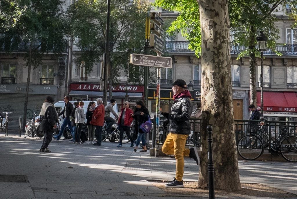 A man standing against a tree in Paris. He wears yellow trousers and leans one leg up.