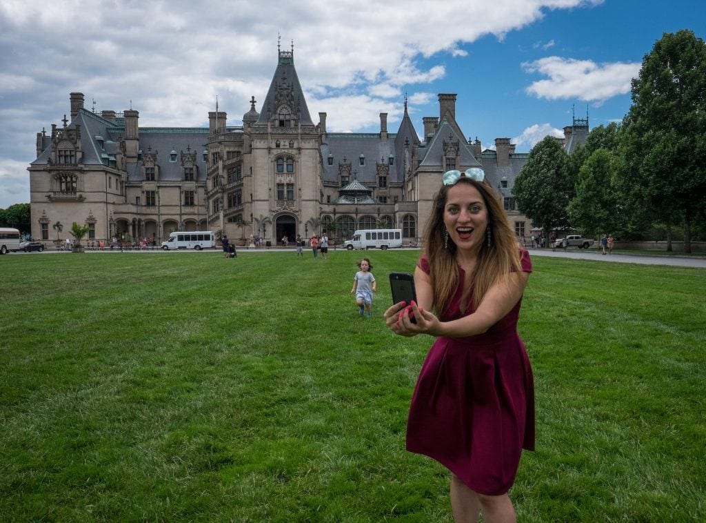 Kate standing and holding her phone while taking a selfie in front of the fancy Biltmore Estate mansion in Asheville, NC.