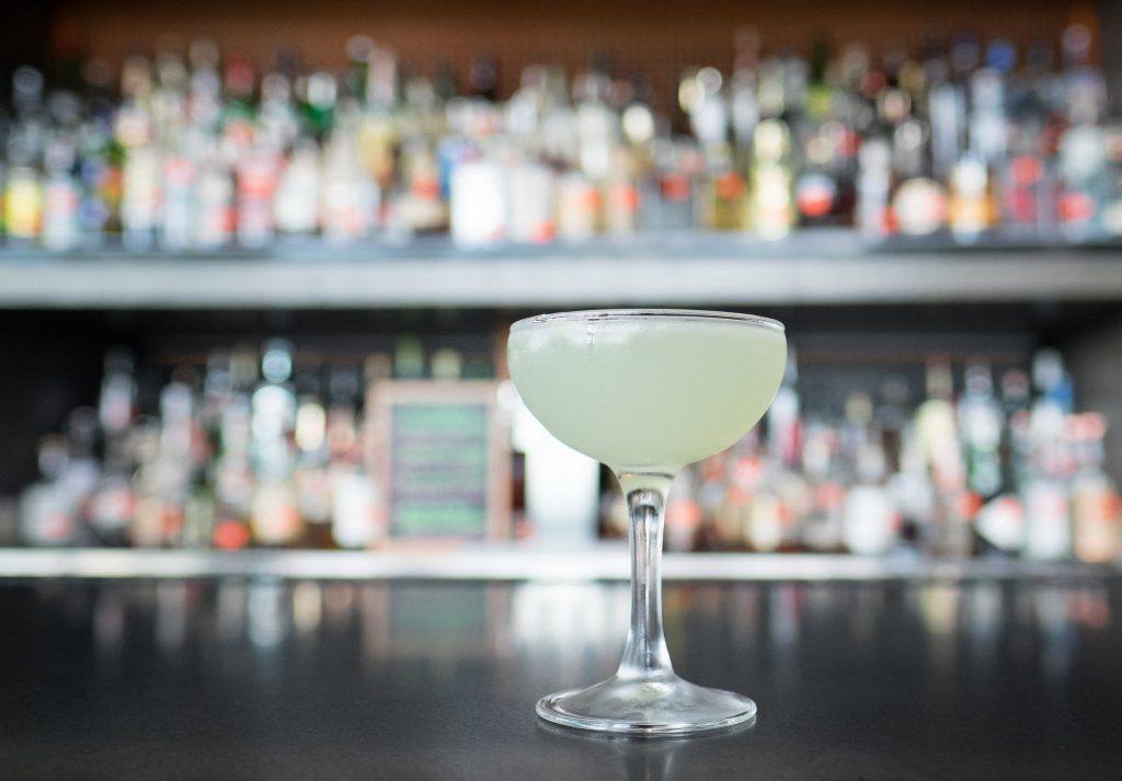A pretty pale yellow cocktail in a margarita class, with bar shelves in the background.