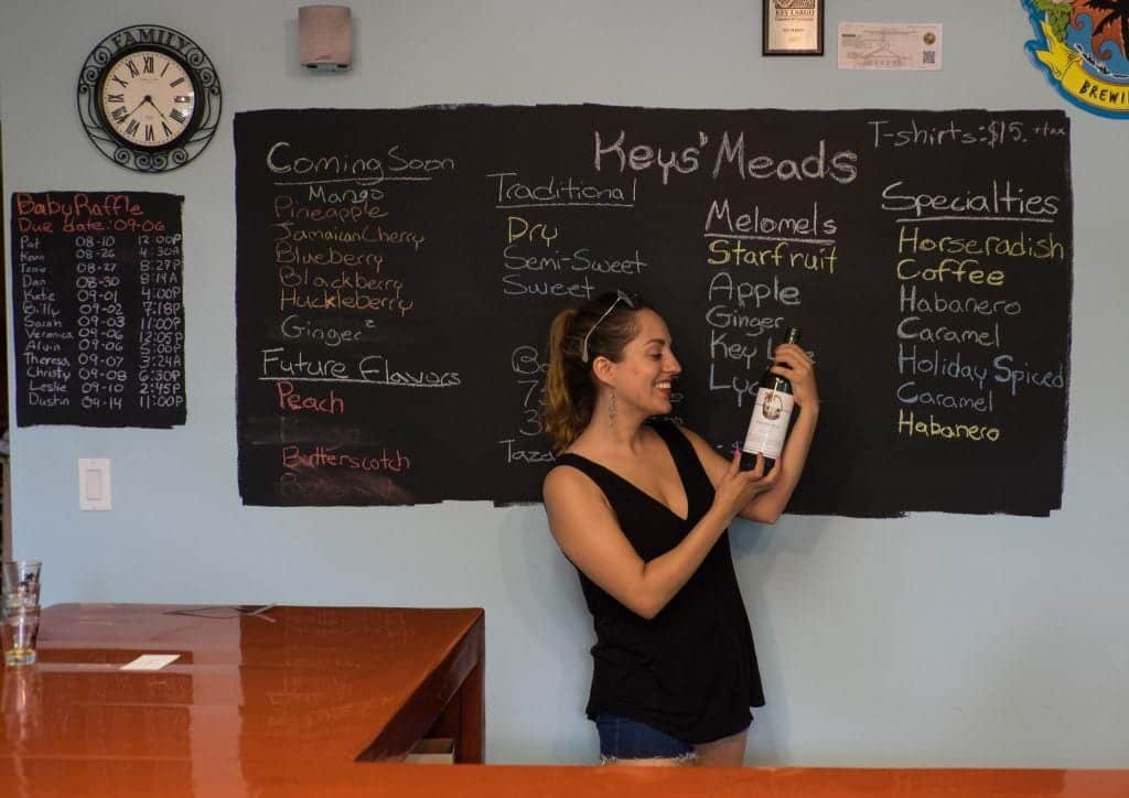 Kate holding a bottle of mead at Keys's Meads, in front of a chalkboard listing the latest flavors.