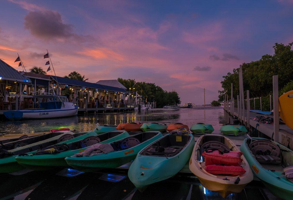 Multicolored kayaks moored on shore at a canal. The sky is a pink, purple and blue sunset.