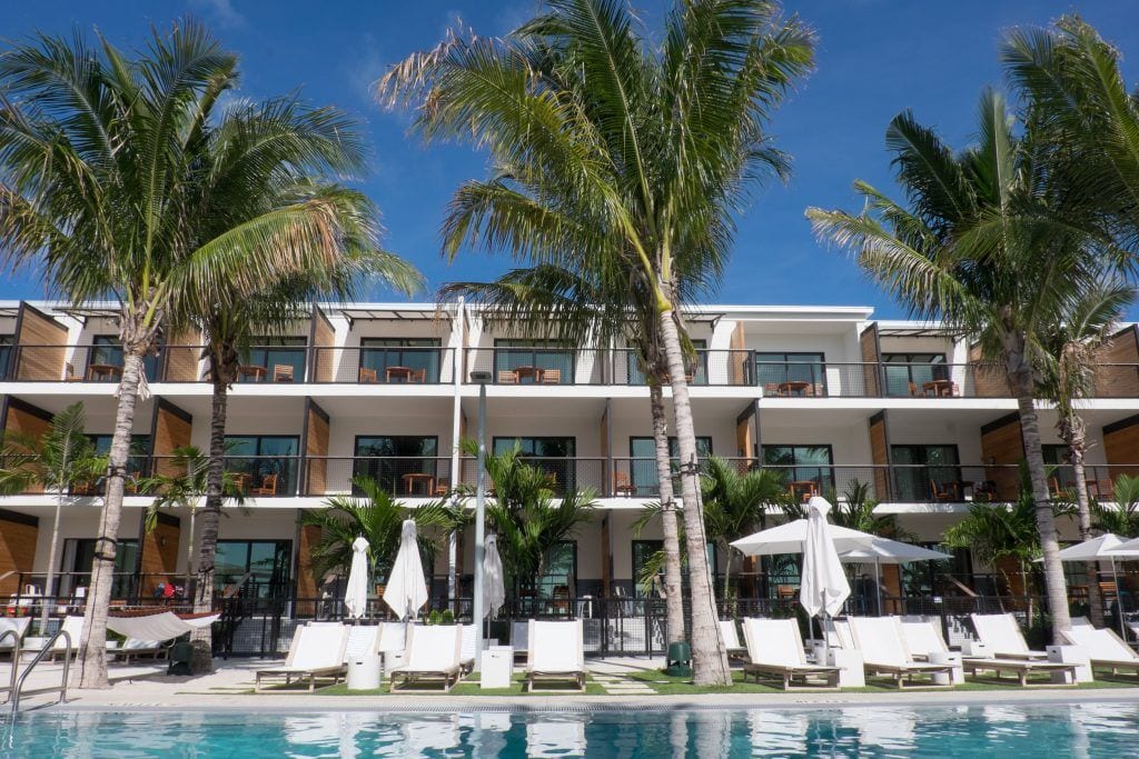 A pool edged with white pool chairs and tall palm trees in front of a bright white modern hotel building.