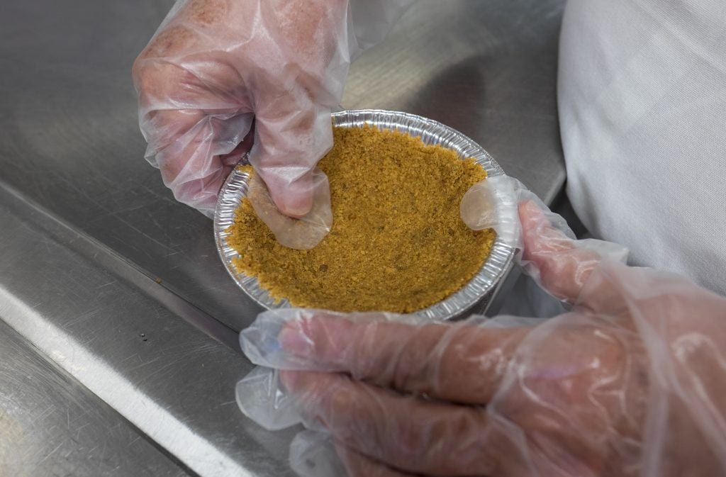 Kate presses a graham cracker crust into a dish in a cooking class, her hands wrapped in plastic gloves.