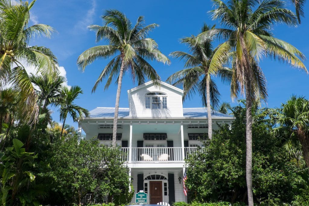 A stately white three-story house with a wraparound porch on the second floor, surrounded by tall palm trees.