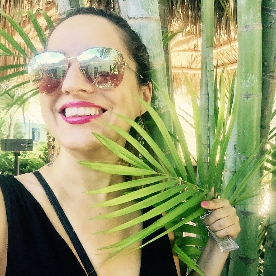 Kate taking a smiling selfie wearing reflective aviator glasses, and holding a palm up to her face.