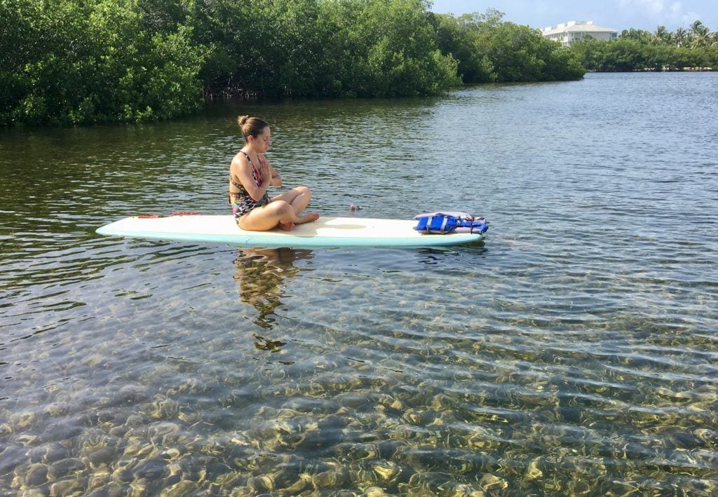 Kate alone doing a sitting yoga pose, her hands together in a prayer position, on a stand-up paddle board on clear turquoise water in the Florida Keys.