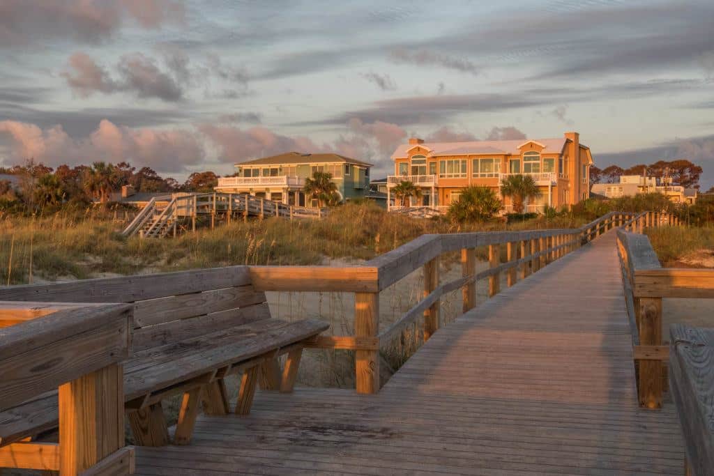 A wooden pathway leading over the sand dunes to large houses, underneath a blue sunrise sky streaked with dark purple clouds.