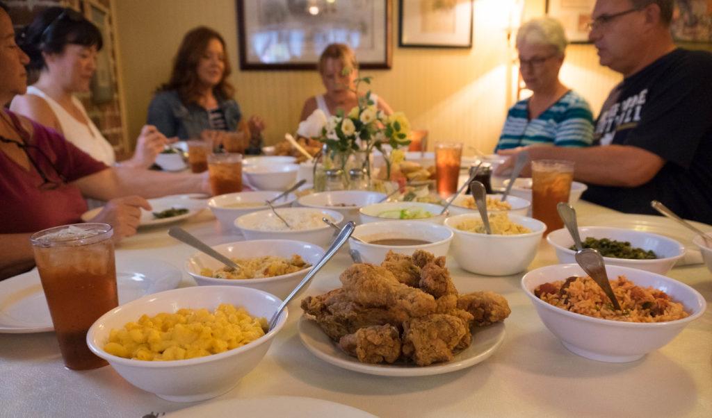 People sitting around a table with lots of bowls of fried chicken, Mac and cheese, an other Southern food.