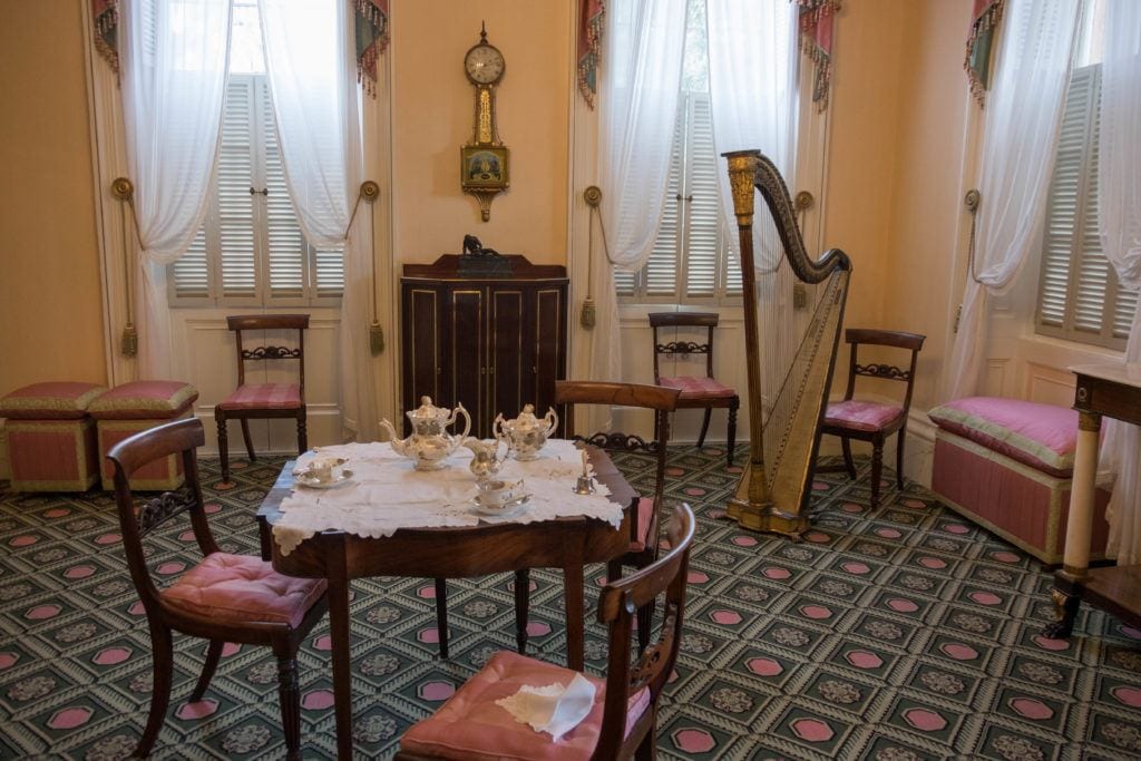 A historic dining room filled with a table and chairs, a harp in one corner, and an old-fashioned clock on the wall.