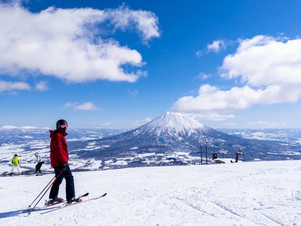 A skier in a red jacket standing on a ski slope, a big snow-covered mountain in the background, underneath a bright blue sky.
