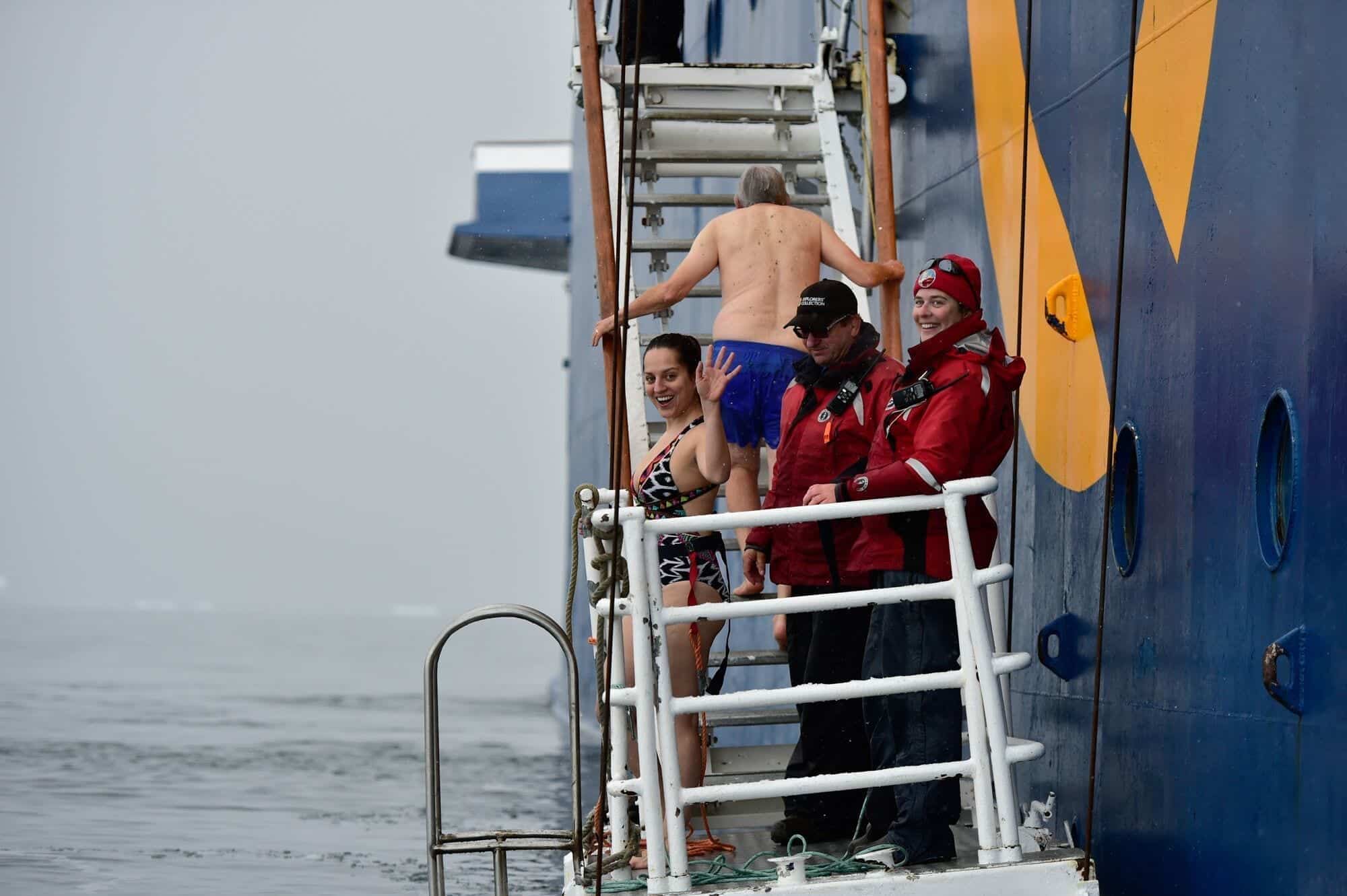 Kate smiling and waving in a patterned one-piece bathing suit while standing on the platform about to jump in the cold Antarctic water, next to two crew members in full winter gear.