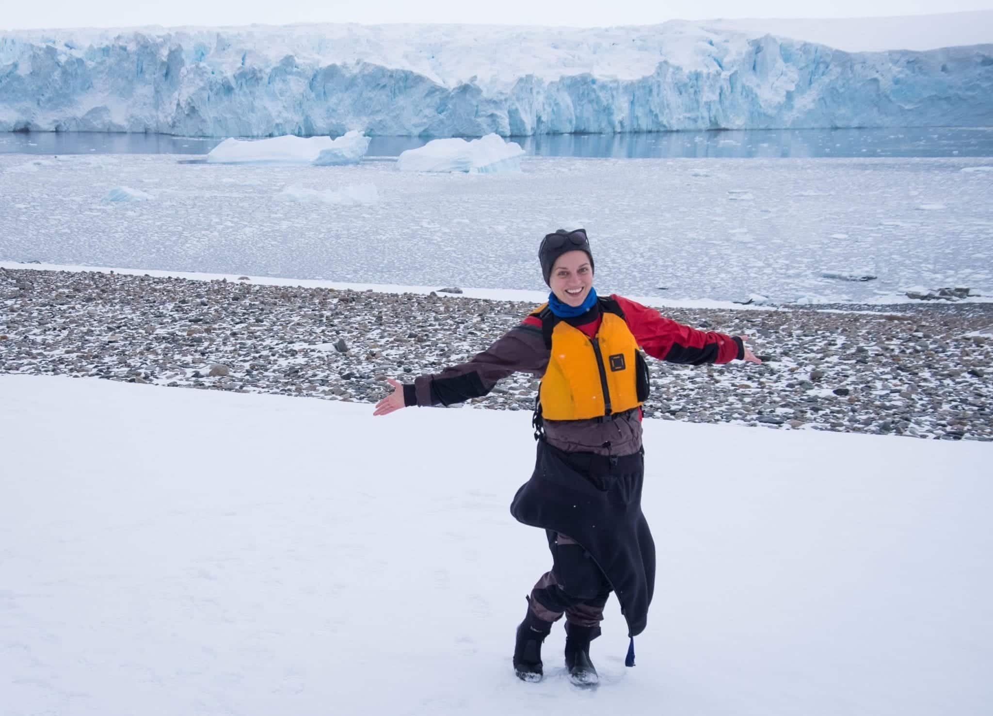 Kate standing in a very snowy and icy part of Antarctica, a huge wall of snow behind her. She holds her hands out and is waring full kayaking gear: a dry suit, life vest, buff, hat, sunglasses, and Kayak skirt.