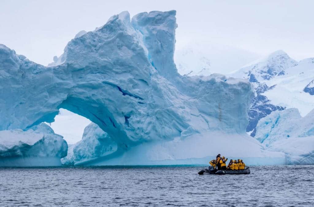 A tiny zodiac boat filled with around 15 passengers in front of a giant ice shape, bluish-white, in Antarctica.