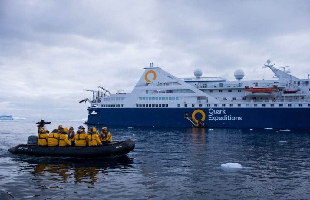 A group of people in yellow jackets on a rubber zodiac boat in front of the Quark Expeditions cruise ship.
