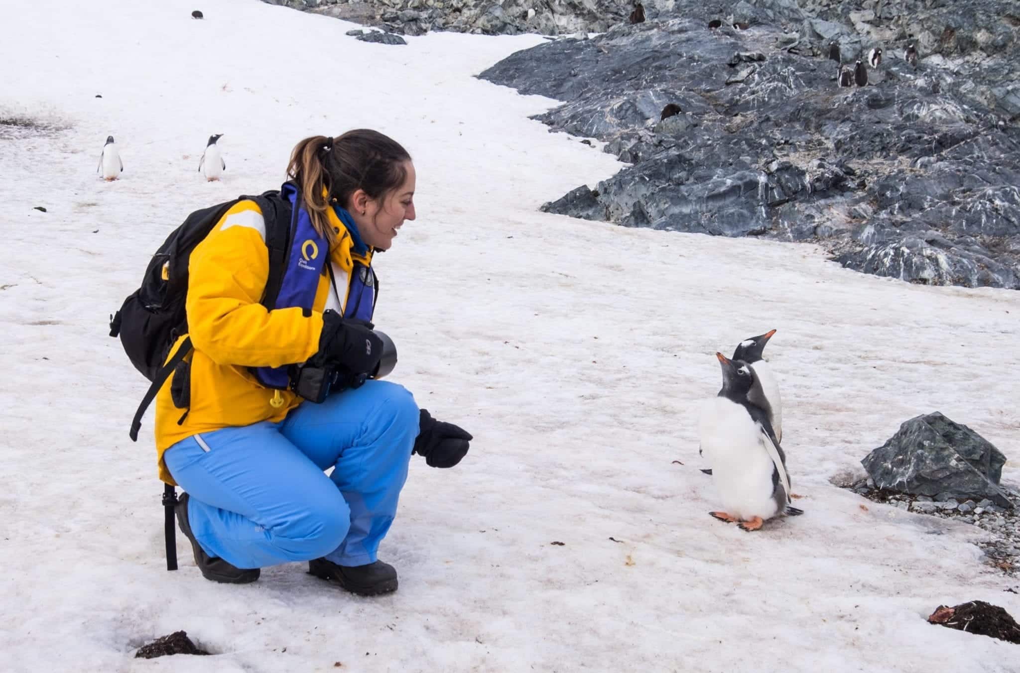 Kate crouching down on the snow in Antarctica, talking to two penguins, their beaks high in the air.