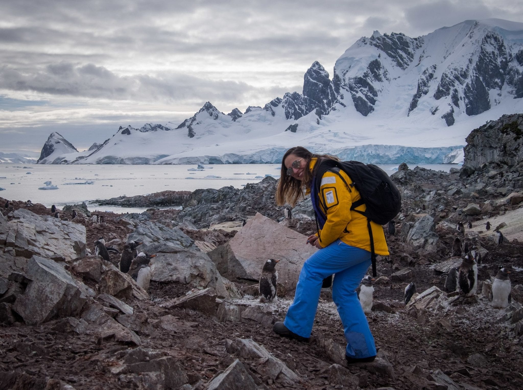 Kate wearing a yellow jacket, blue pants, and sunglasses and stepping on a rocky pile next to a penguin, tall snowy mountains behind her.