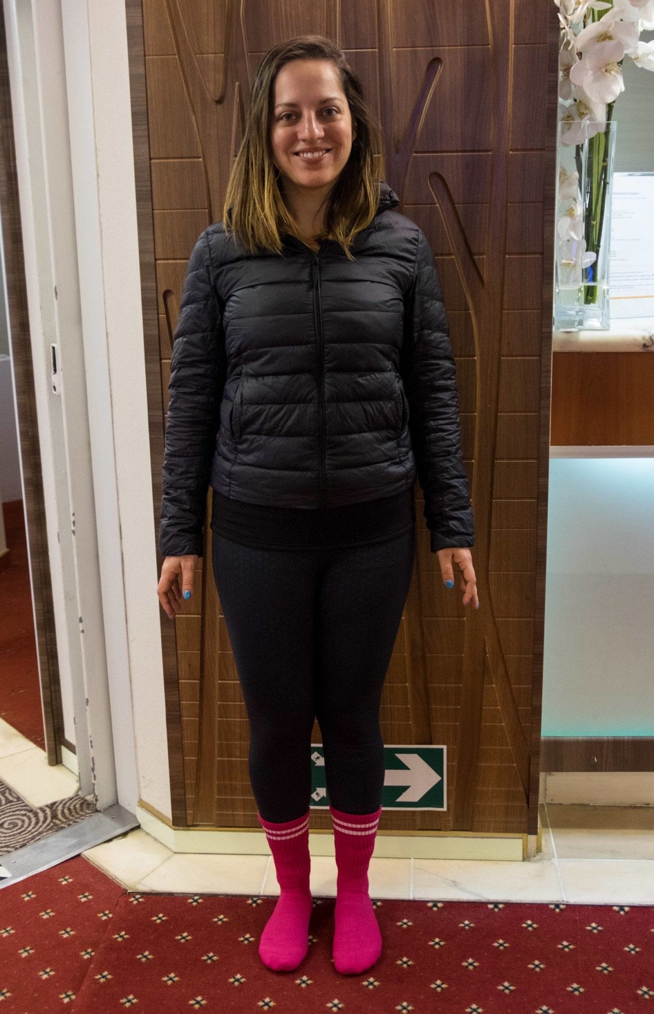 Kate standing in the same position, now with a black puffer jacket, black leggings, and bright pink thick socks on top of what she was wearing before.