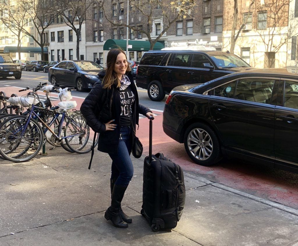 Kate standing on the street in New York, wearing jeans and a black jacket, holding a large black rolling suitcase.