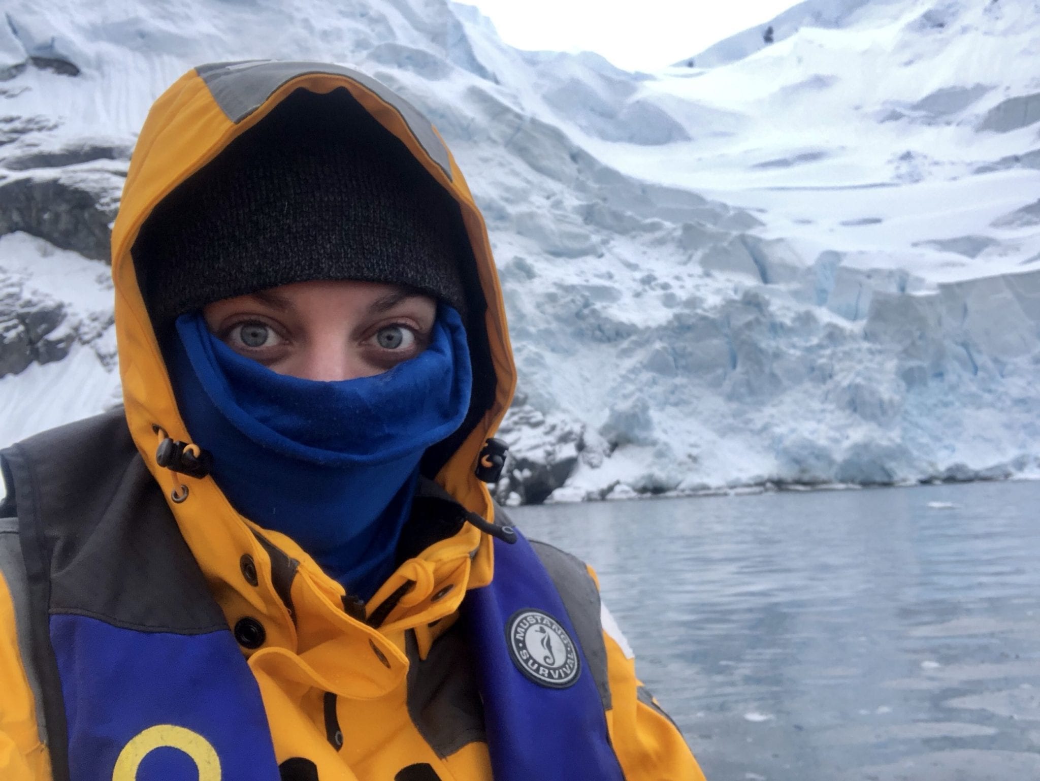 Kate taking a selfie in front of a big snowy wall. She has her yellow hood up, black hat on, and a bright blue buff covering her face beneath the eyes.