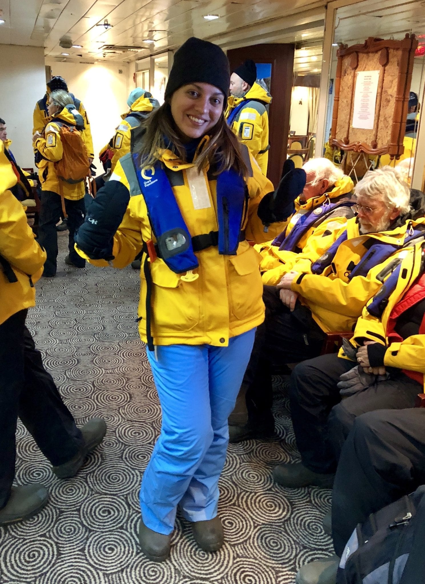 Kate standing in a crowd about to disembark the boat, wearing a bright yellow coat, blue life vest, black beanie, black mittens, baby blue snow pants, and dark green boots.