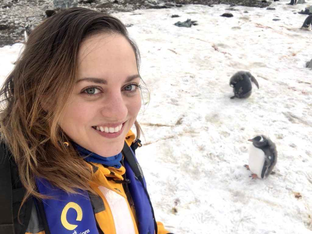 Kate taking a smiling selfie next to two penguins in the snow on the ground.
