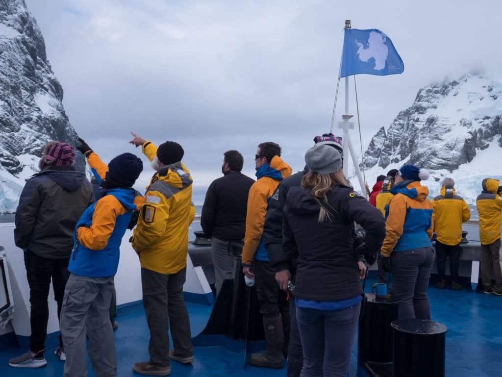 A group of people standing on board an Antarctica ship and pointing at snow-covered cliffs.