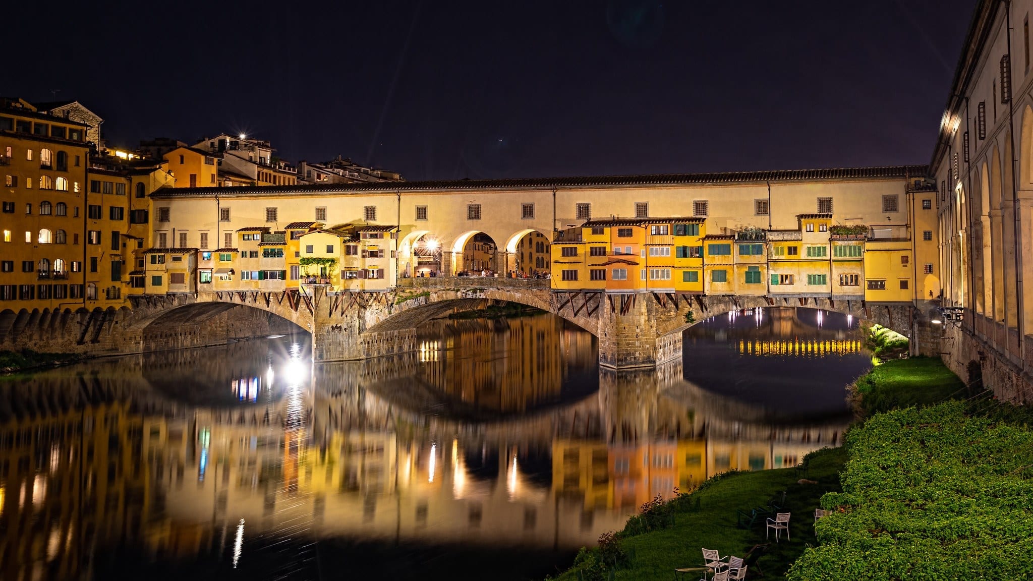 A bridge in Florence lit up at night with yellow buildings in the background, reflected into the water.