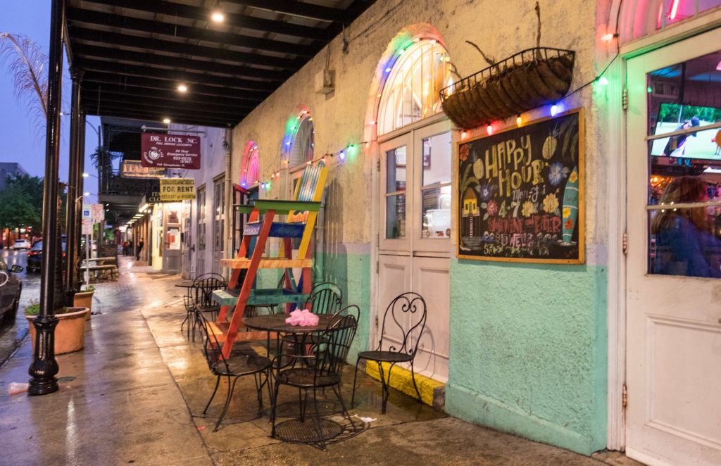 A New Orleans cafe on the street at night, colored lights dangling outside, people sitting on wrought iron chairs and enjoying cocktails.