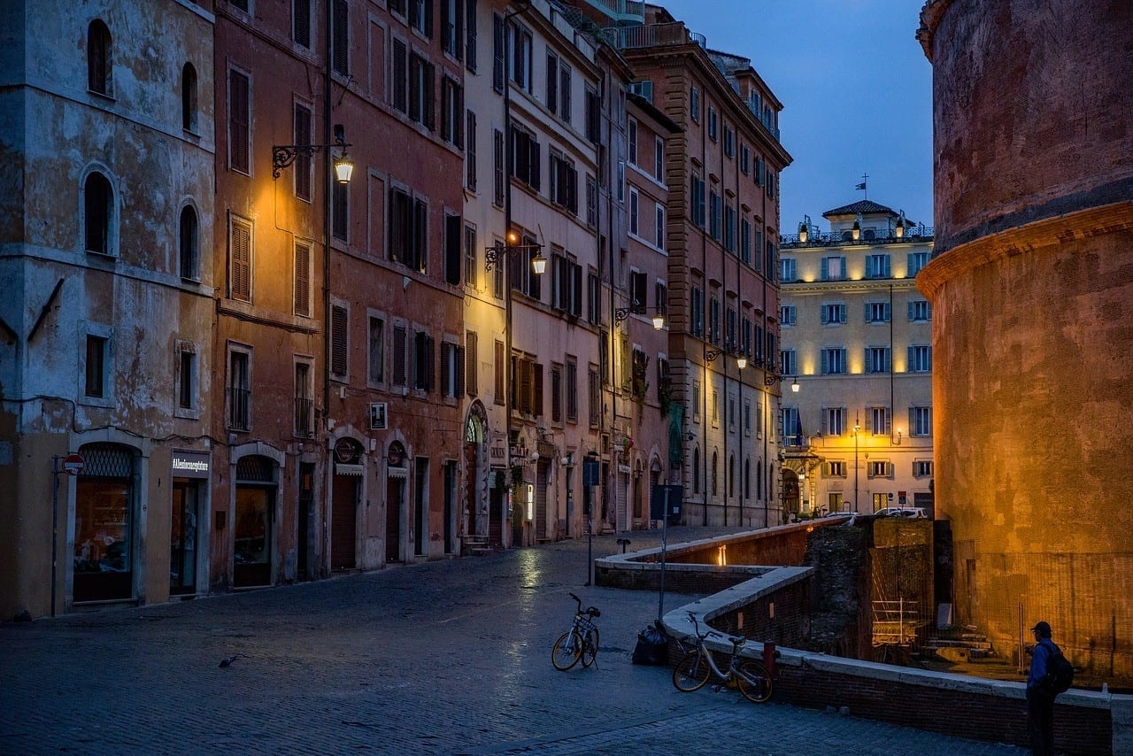 A small alleyway in Rome at dusk, brightly colored buildings lit by yellow streetlamp.