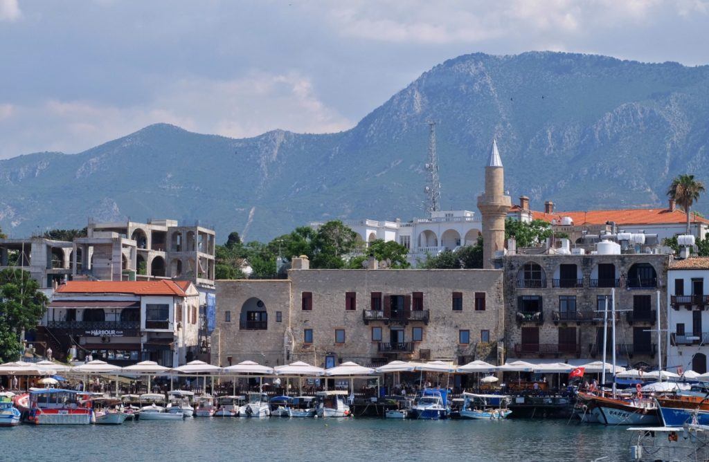 A coastal town in Cyprus with umbrella-topped cafe tables at the water's edge, and a mosque's minaret, in front of a mountainous backdrop.