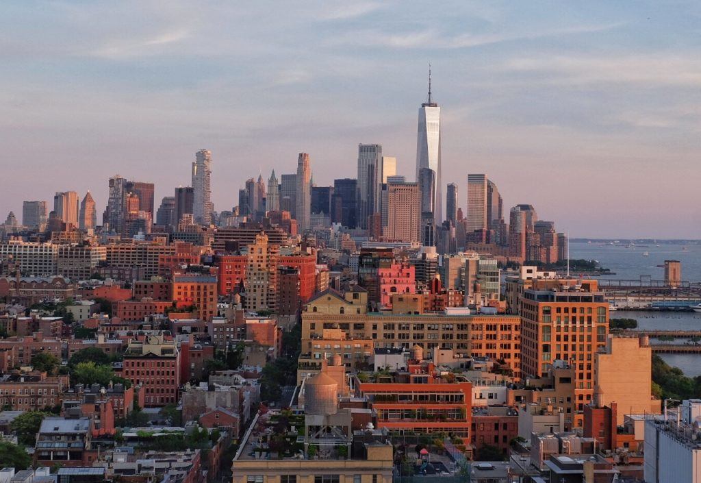 The New York skyline as seen from Hell's Kitchen, the buildings brown and silver underneath a blue and pink sunset.