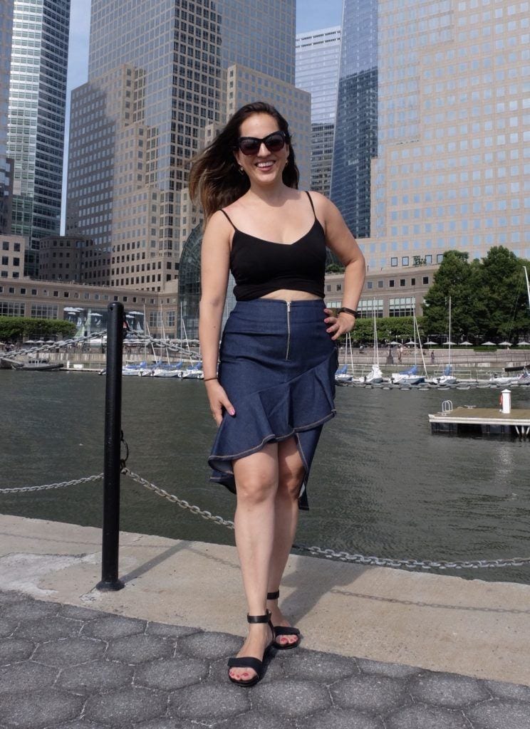 Kate standing in a black crop top and blue denim skirt in front of modern skyscrapers in lower Manhattan.