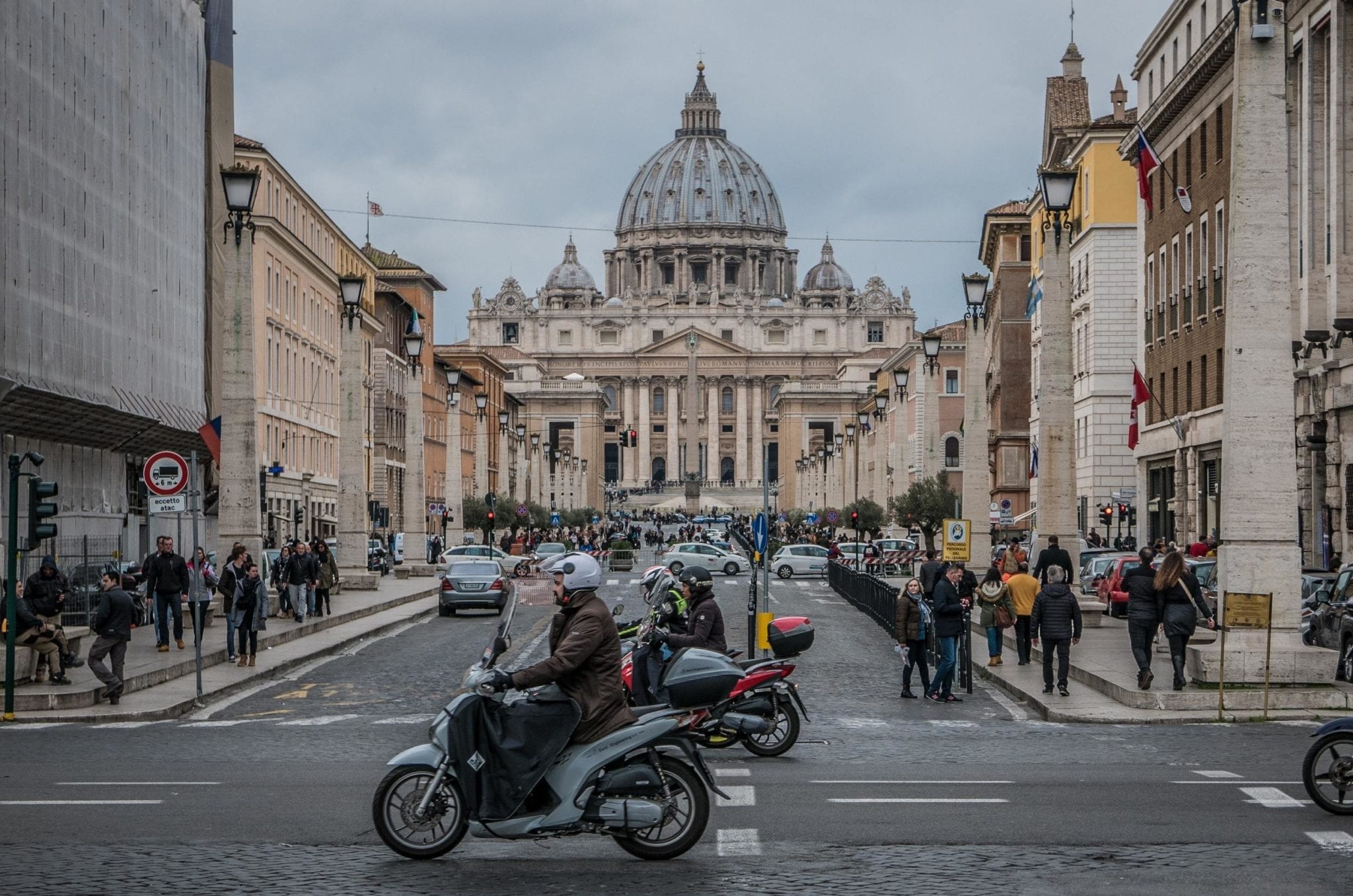 A street view of Rome with a man on a motorbike in front of the large dome of St. Peter's Basilica.