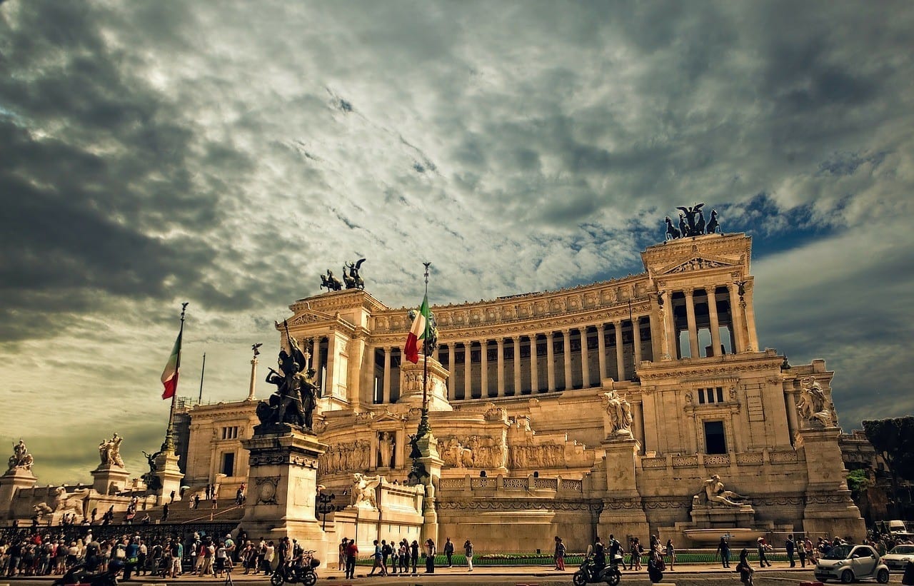 Vittorio Emanuele, the "wedding cake" building of Rome with large columns and Italian flags flying, bright yellow at dusk beneath a cloudy sky.