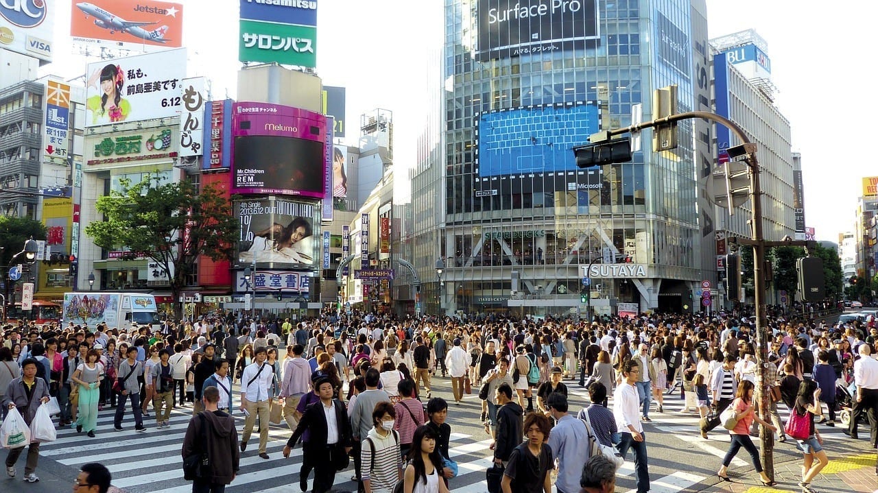 Hundreds of people cross the enormous Shibuya Crossing in Tokyo as tall buildings covered with screens and advertisements surround them.