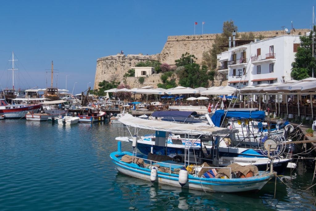 The waterfront of Girne, Cyprus: you see calm bright blue water, colorful wooden boats in the foreground and a tall stone fortress in the background, all underneath a bright blue sky.