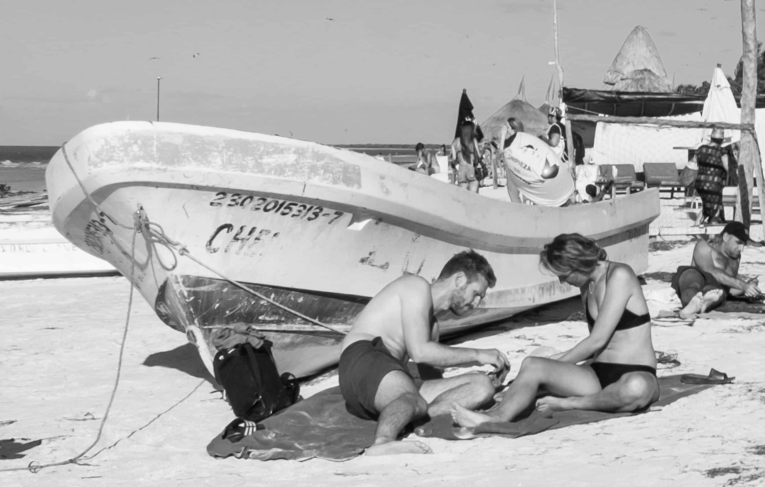 Black and white photo of a couple sitting on the beach beside a boat.