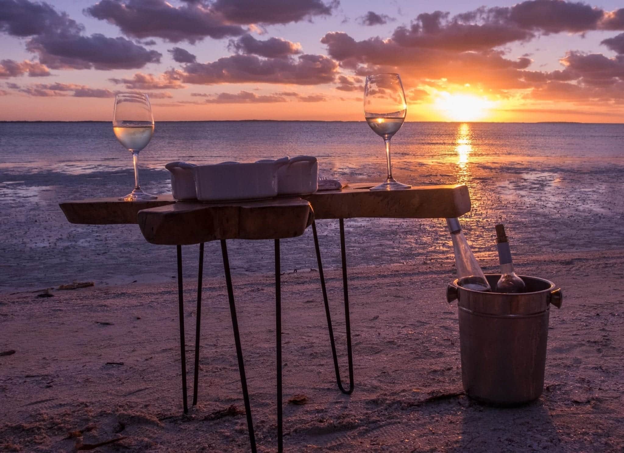Table on the beach at sunset with two wine glasses and an ice bucket with two bottles of wine.