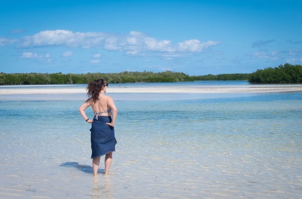 Kate wearing a long denim skirt and bathing suit top, facing outwards while standing in ankle-deep clear blue water in Holbox.