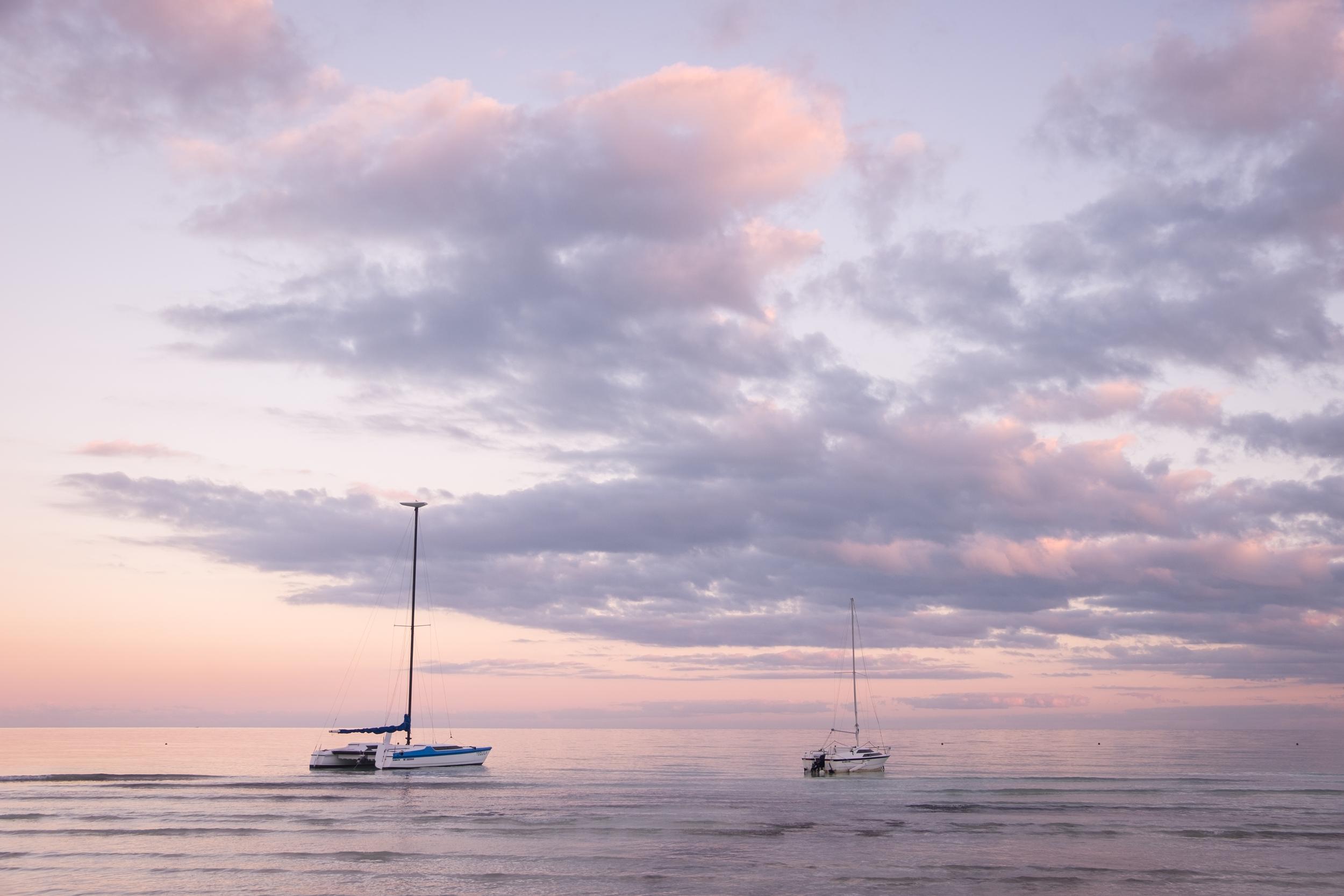 Two sailboats in the water with pink skies and clouds.