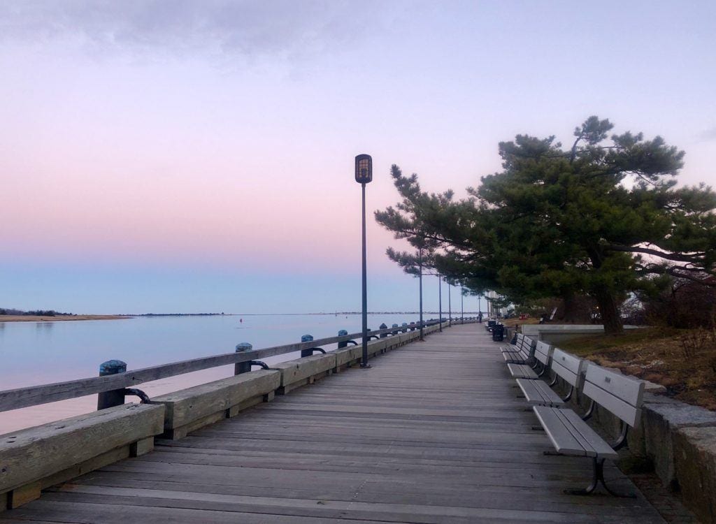 A wide wooden pathway along the water in Newburyport, Massachusetts, lined with benches. There is a pink and purple sunset and it reflects in the water.