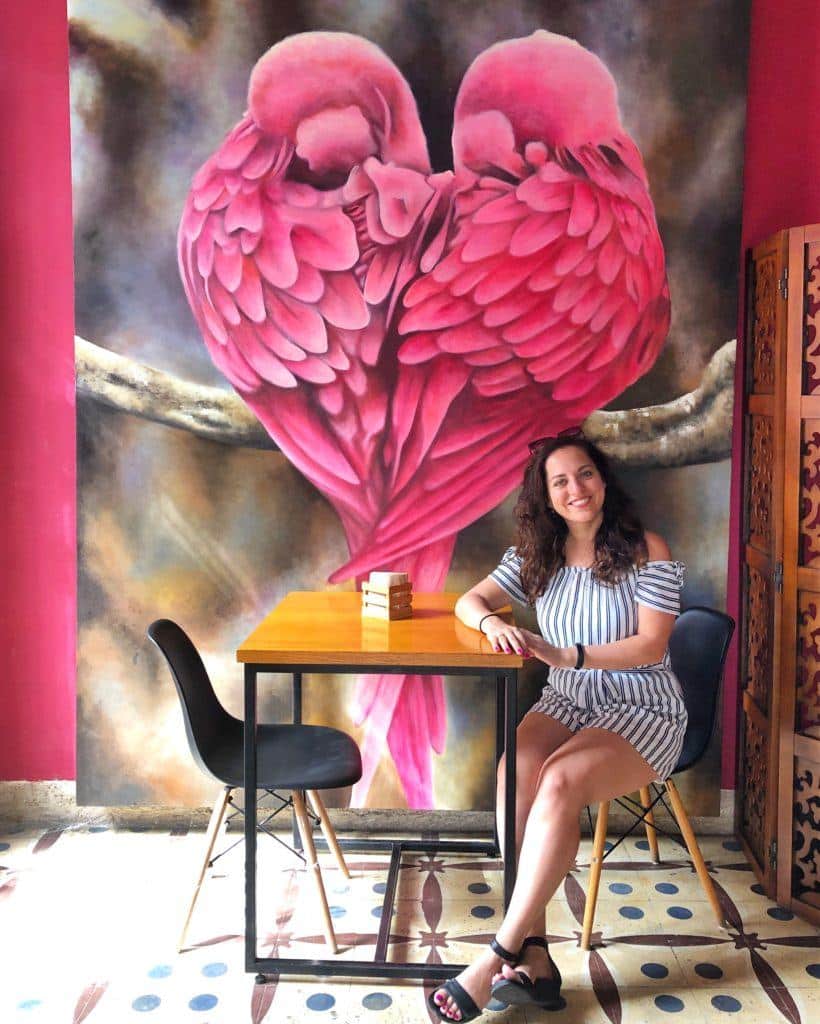 Kate sits at a cafe in front of a mural of two pink birds shaped like a heart, an open seat across from her.