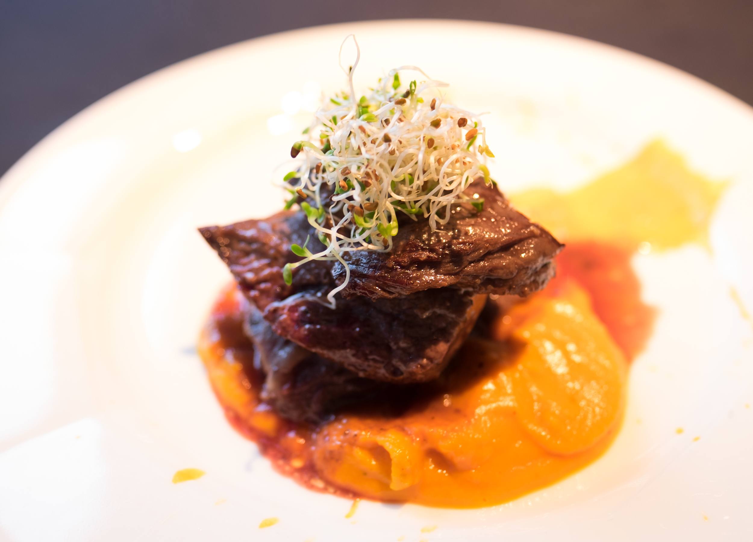 Three little pieces of steak on a bright orange sauce, topped with sprouts.