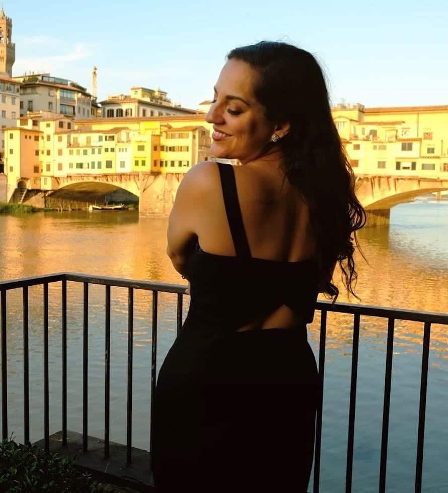 Kate wears a long black sleeveless dress and is facing her body away from the camera but turning back toward it with her face, smiling with her eyes closed. Behind her is the Ponte Vecchio, the old bridge of Florence covered with jewelry stores, bathed in golden light.