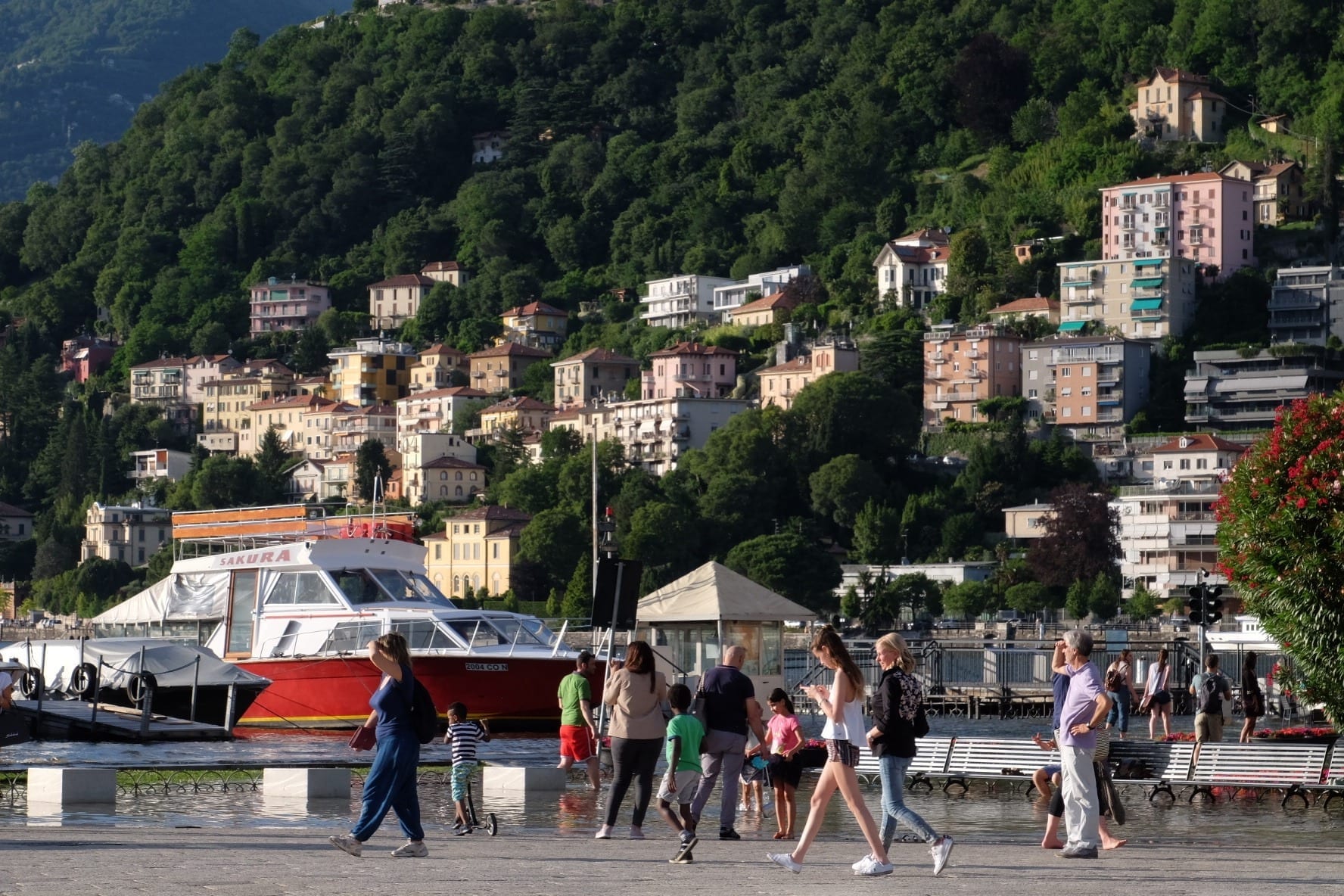 Several people walk across a piazza in Como, Italy. In the background is the lake and several houses built on a green mountainside leading into the lake.