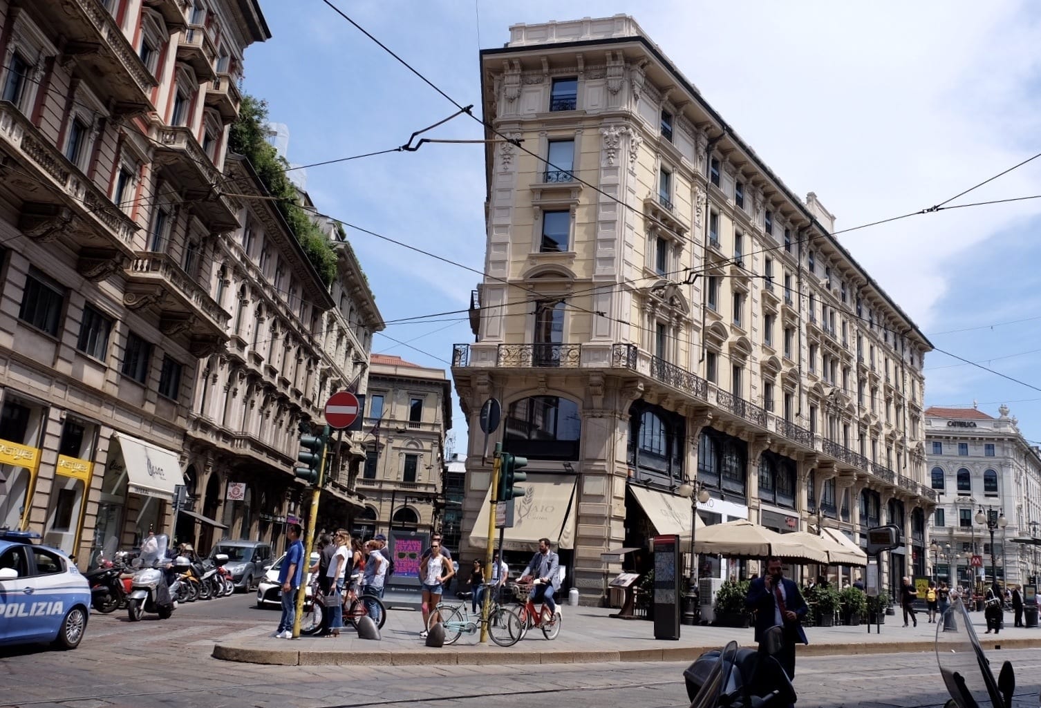 Milan street scene: On a block that juts out triangularly into the street, several people wait for the walk signal at a stoplight. The surrounding buildings are gray with ornate balconies for each window. On the ground floor is a cafe with white awnings.