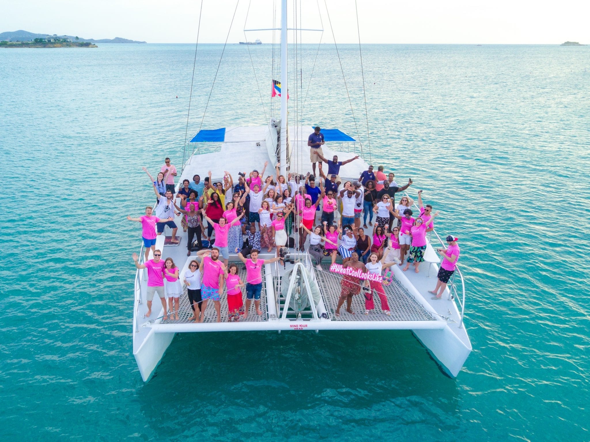 In a drone shot, a few dozen people, some wearing bright pink t-shirts, pose with their arms in the air on a large catamaran in Antigua. The sea surrounding them is turquoise.