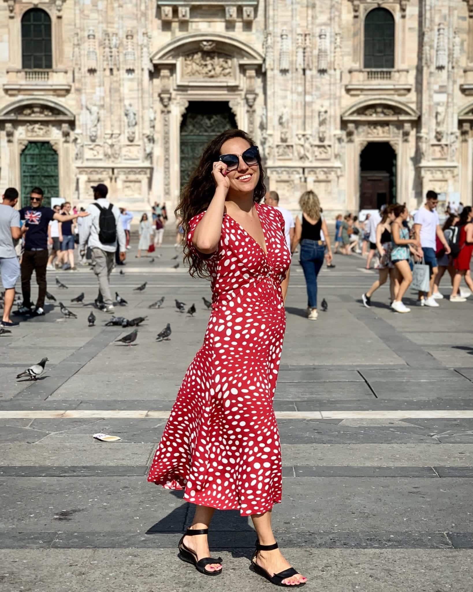 Kate wears a long red short-sleeved dress covered with white polka dots of various sizes and black sandals. She wears black sunglasses and poses as if about to take them off with one hand. She is standing in front of Milan's Duomo and in the background you can see pigeons and people taking pictures in front of it.